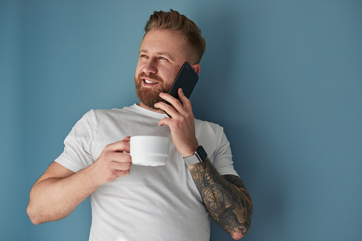 Glad bearded guy with cup of fresh coffee smiling and looking away while answering phone call against blue wall