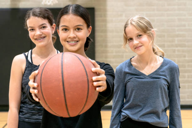 Female Students Playing Basketball Three female elementary students pose for a portrait while in gym class. b They are each dressed comfortably in athletic wear and the girl in the middle is holding out a basketball. youth basketball indoor stock pictures, royalty-free photos & images
