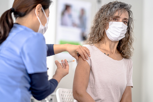 A middle-aged woman sits up tall and still as a female nurse of Asian decent administers her COVID-19 vaccination.  She is dressed casually and looking away as the vaccine is administered.  Both are wearing medical masks to protect them from the virus.