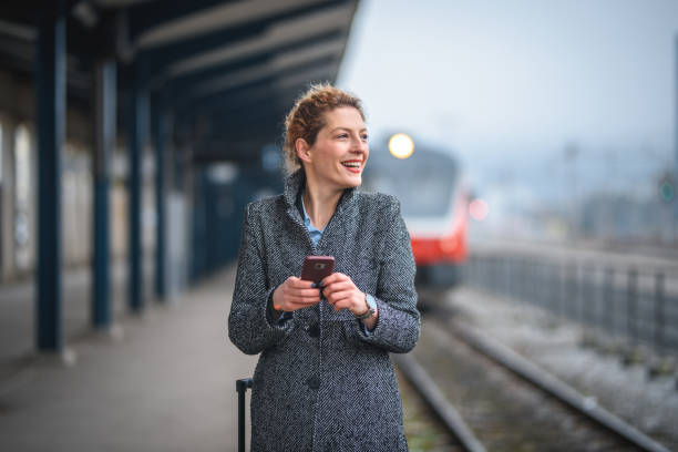 Young business woman at railway station, elegantly dressed, walking along the platform and pulling a small suitcase, checking directions on her mobile phone and smiling. Full length image.