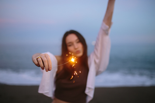 Let's sparkle things up. Carefree young woman holding a sparkling light in her hand while standing at the beach in the evening. Fun-loving young woman celebrating with bengal lights.