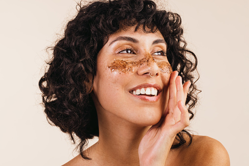 Confident young woman looking away with a smile while applying coffee scrub on her face. Happy young woman exfoliating her facial skin using a cleansing coffee mask.