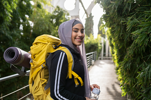 Pretty young Muslim woman hiker smiling while looking at the camera in nature with a backpack. - woman traveler -