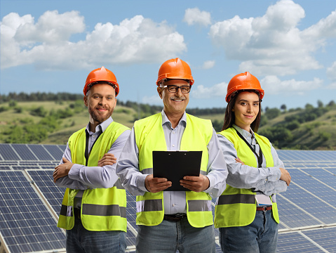 Team of young and mature engineers with safety vests on a solar farm smiling at camera