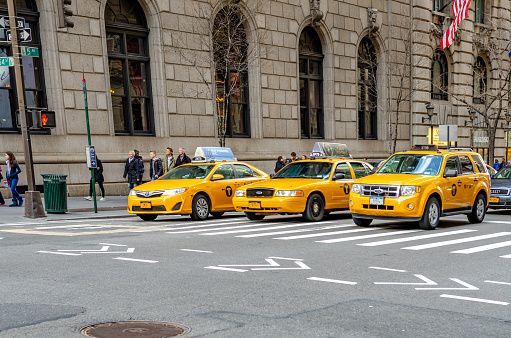 Yellow Taxi cabs, three different types of cars, waiting for the traffic light to turn green, crosswalk in front, Fifth Avenue, with People walking on sidewalk, Manhattan, New York City during winter day, horizontal