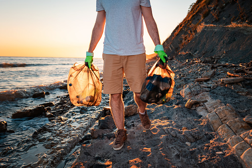 Close up of volunteer walking along at the wild beach holding two plastic bags. In the background, the ocean and the sunset. The concept of cleaning the beach from pollution.