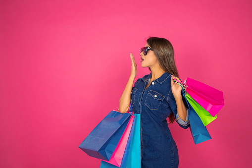Beautiful girl with denim dress and sunglasses carrying shopping bags on pink background. Concept of shopping, summer sale. Online shopping concept, copy space provided. The scene is situated in controlled studio environment in front of pink background. Photo is taken with SONY A7III camera.