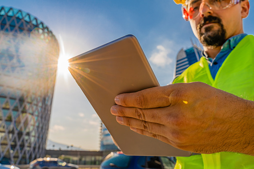 Close-up of young engineer or architect with protective workwear using digital tablet  in front of modern business buildings and construction site. Low angle view and lens flare. The model is young male adult caucasian ethnicity who can be engineer,  architect or construction worker. The photo is shot with Sony A7III camera.