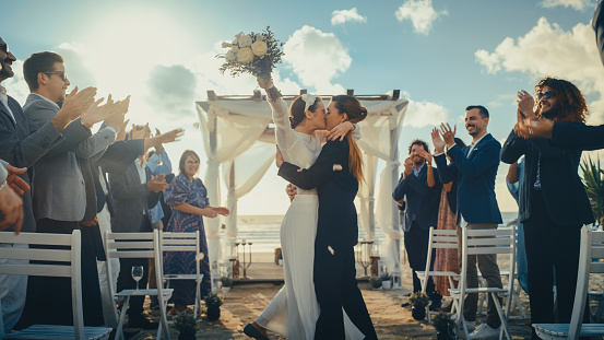 Atractiva pareja femenina queer camina por el pasillo en la ceremonia de boda al aire libre cerca del océano. Dos mujeres lesbianas enamoradas comparten felicidad con diversos amigos multiétnicos. Lindos objetivos de relación LGBTQ. photo