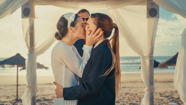 beau couple queer féminin échanger des bagues et s’embrasser lors d’une cérémonie de mariage en plein air près d’une mer. deux femmes lesbiennes amoureuses partagent leur grand jour avec diverses amies multiethniques. objectifs de la relation lg - homosexual wedding married engagement photos et images de collection