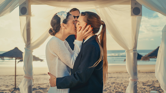 Beautiful Female Queer Couple Exchange Rings and Kiss at Outdoors Wedding Ceremony Near a Sea. Two Lesbian Women in Love Share Their Big Day with Diverse Multiethnic Friends. LGBTQ Relationship Goals.