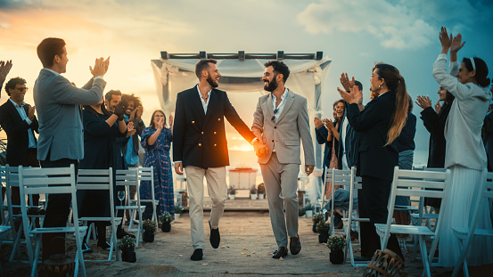 Handsome Gay Couple Walking Up the Aisle at Outdoors Wedding Ceremony Venue Near Sea. Two Happy Men in Love Share Their Big Day with Diverse Multiethnic Friends. Authentic LGBTQ Relationship Goals.