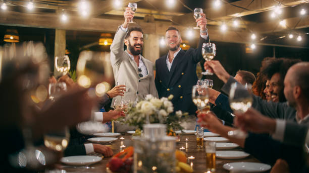 big dinner party with a crowd of multiethnic diverse friends celebrating at a restaurant. handsome happy hosts propose a toast and raise wine glasses while sitting at a table in the evening. - wedding champagne table wedding reception imagens e fotografias de stock