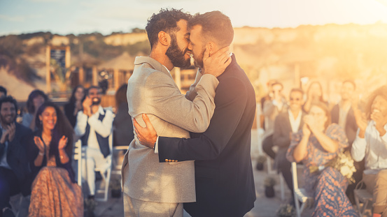 Hermosa pareja gay intercambia anillos y besos en el lugar de la ceremonia de boda al aire libre cerca del mar. Dos hombres felices enamorados comparten su gran día con diversos amigos multiétnicos. Objetivos de relación LGBTQ. photo