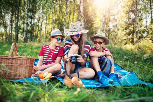 Three kids having picnic sitting on blanket in forest. They are looking at the photos  taken with the mirrorless camera.Nikon D850