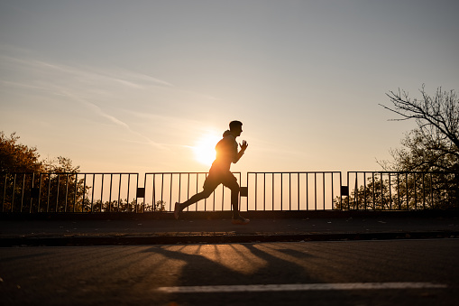 Wide angle side view of a silhouette of a male sportsperson doing his daily running routine outdoors with a sun setting on the other side