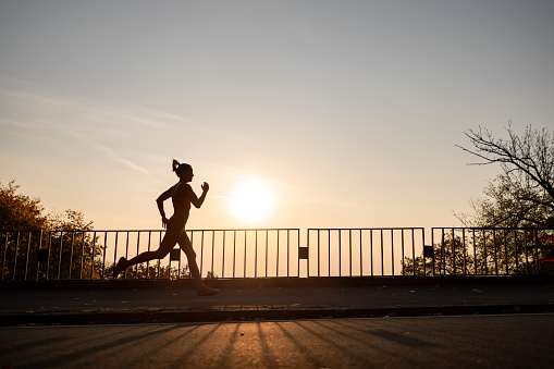 Wide angle side view of a silhouette of a sportswoman doing her daily running routine outdoors with a sun setting on the other side