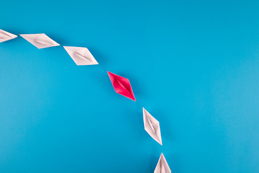 Group of white paper boats and one red paper boat going same direction on blue background