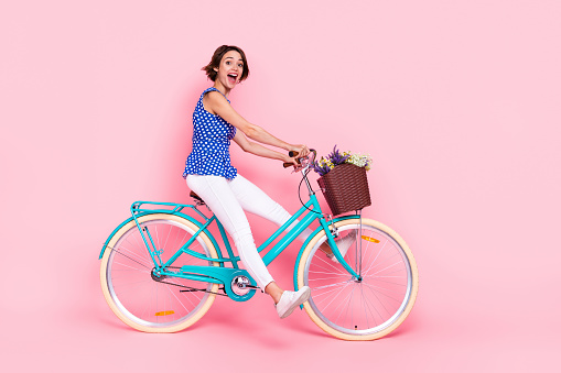 Full length profile photo of impressed millennial lady ride bicycle wear blouse pants sneakers isolated on pink background.