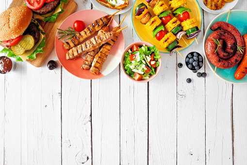 Summer BBQ or picnic food top border. Variety of burgers, grilled meat, vegetables, fruits, salad and potatoes. Overhead view on a white wood background. Copy space.