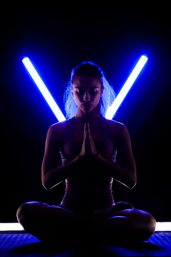 Fit woman practicing yoga poses. Silhouette girl doing exercise in studio against black background with v shaped neon blue or purple led tube light.