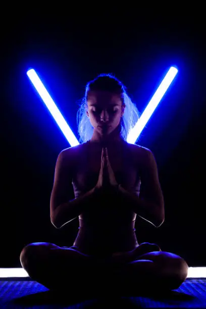 Photo of Fit woman practicing yoga poses. Silhouette girl doing exercise in studio against black background with v shaped neon blue or purple led tube light.