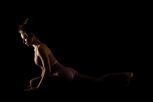 Fit woman practicing yoga poses. Side lit half silhouette girl doing exercise in studio against black background.