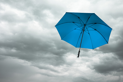 A Blue umbrella fly over the dark grey cloudy sky with stormwind in the rainy season, depth of field. Parasol blown away by heavy windy weather into dramatic cloudscape, overcast clouds background.