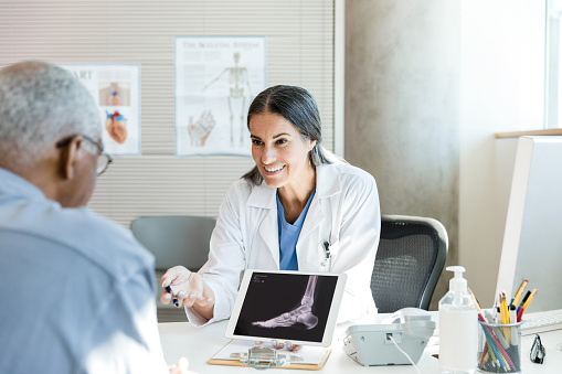 A mid adult female orthopedic surgeon smiles encouragingly while discussing the x-ray results with the unrecognizable senior adult patient.  She is using a digital tablet to display the image of his foot.