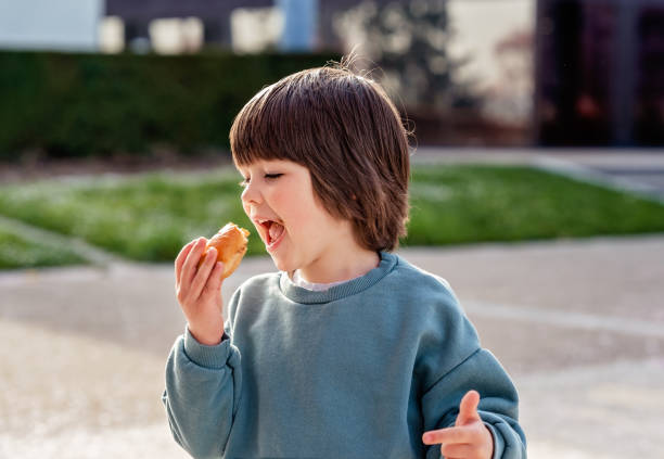 Hungry toddler boy enjoying snack taking a bite while walking outside in park at spring sunny day. stock photo