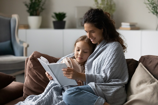 Smiling caring mother with little daughter reading book together, happy mom with adorable girl child wrapped in warm blanket sitting relaxing on comfortable couch, engaged in educational activity