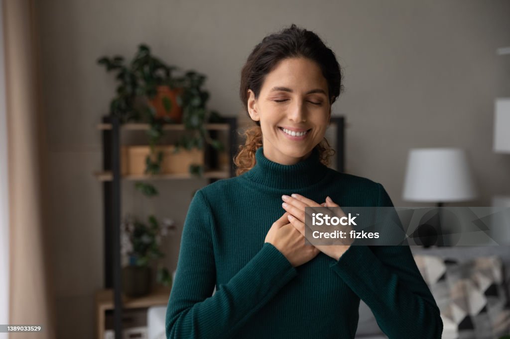 Happy Latin woman hold hands on heart feeling grateful Smiling young Hispanic woman hold hands at heart chest feel grateful and thankful. Happy millennial Latino female believer show love and care support pray or mediate. Faith, superstition concept. Chest - Torso Stock Photo