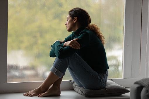 Pensive sad millennial Hispanic woman sit on windowsill at home look in window distance thinking dreaming. Thoughtful young Latino female loner or outcast feel lonely relax rest indoors on rainy day.