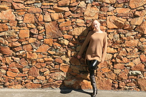 A Caucasian model leaning against a brown stone wall. She is wearing a brown sweater with black, shiny pants and boots.