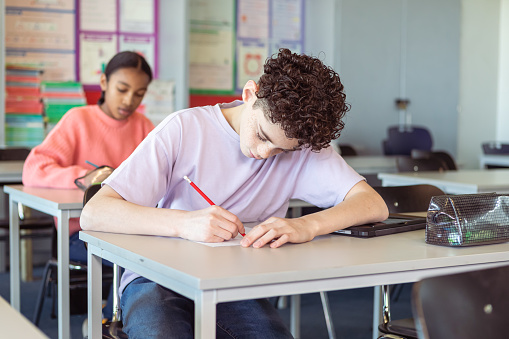 A diverse group of adolescent boys and girls sit at their school desks and take a test during class.