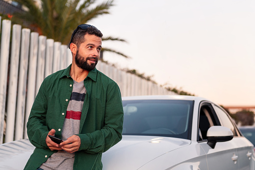 attractive young bearded man sitting on the hood of his car with mobile phone in hand, leisure and communication technology concept, copy space for text