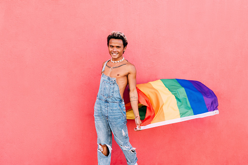 Happy male in denim casuals standing with LGBT flag at pink wall