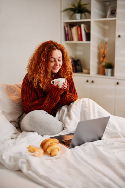 istock A ginger girl sits on the bed in the morning, drinking her morning coffee and using her laptop. 1389094607