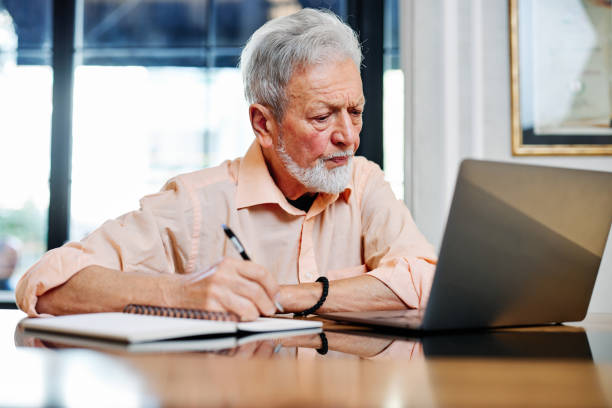 istock A senior student is sitting at home and writing down notes in a notebook while following an online lecture on the laptop. 1389096426
