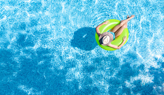 Active young girl in swimming pool aerial top view from above, teenager relaxes and swims on inflatable ring donut and has fun in water on family vacation, tropical holiday resort