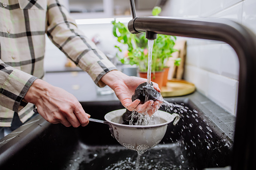 A woman cleaning shungite stones in sieve with pouring water in sink.