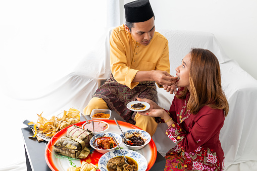 Young Muslim couple celebrates end of Ramadan, with husband feeding wife, symbolic of love, with traditional food cuisines from a circular tray, sitting on white settee, white background. Its the Ramadan Mubarak, fasting, Ramadan Kareem season.