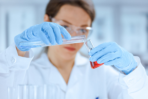 A young white female chemist in a laboratory coat and protective gloves mixes colored liquids in a laboratory flask. The focus is on a flask and a tube on the foreground