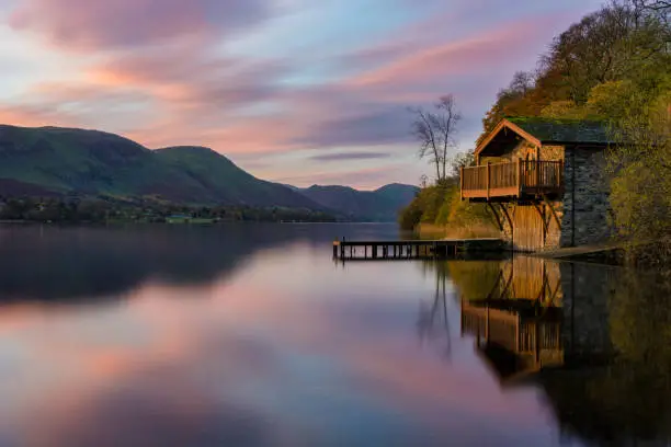 Pink and purple vibrant sunrise with dawn light hitting boathouse on calm lake with reflections in the Lake District.