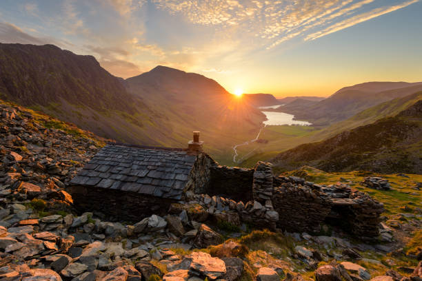 Beautiful Vibrant Sunset In The English Lake District With Stone Mountain Hut. A scenic view seen from Warnscale Bothy, overlooking Buttermere and Crummock Water in the Lake District, UK. Golden light from the setting sun can be seen illuminating the rocks in the foreground that surround this iconic landmark. english lake district stock pictures, royalty-free photos & images
