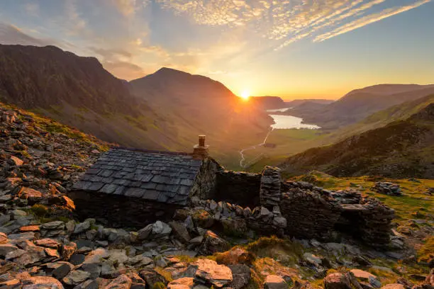 A scenic view seen from Warnscale Bothy, overlooking Buttermere and Crummock Water in the Lake District, UK. Golden light from the setting sun can be seen illuminating the rocks in the foreground that surround this iconic landmark.