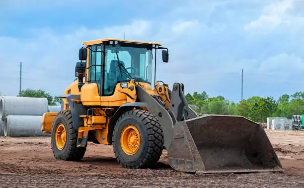 construction site with wheel loader in foreground, sewer pipe and tree line in background, sunny day, no people, machinery on dirt with bucket down on ground, blue sky with some clouds