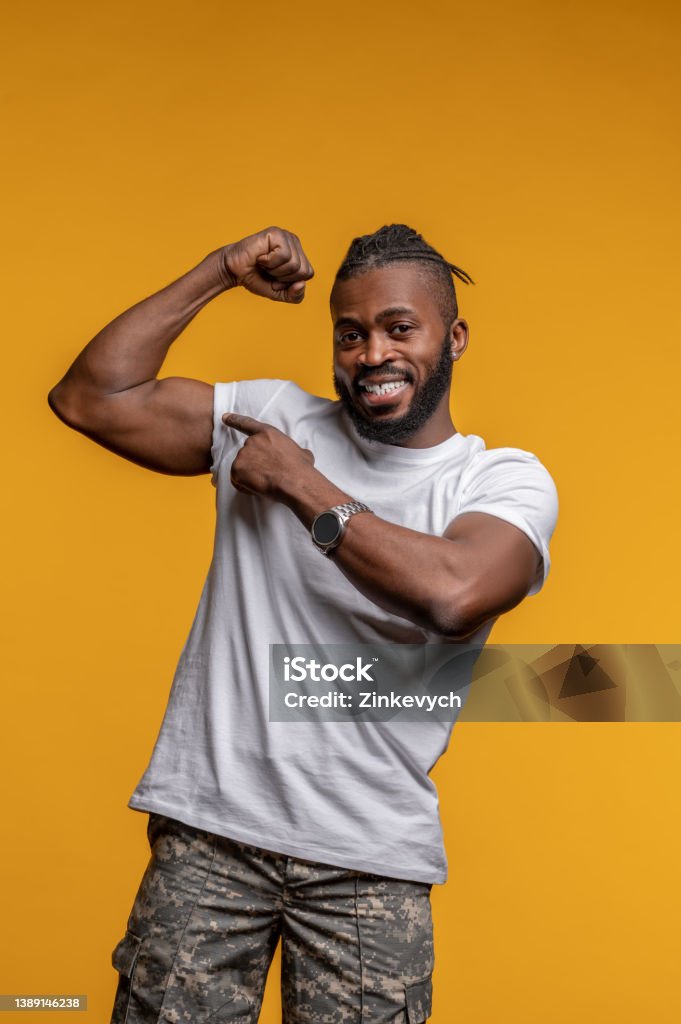 Pleased young man demonstrating his physical strength Smiling happy cute athletic African American male pointing at his biceps on the upper arm Bicep Stock Photo