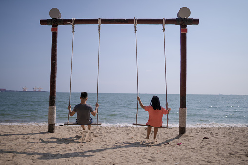 2 Asian Chinese brother and sister swings on beach chair, above sea