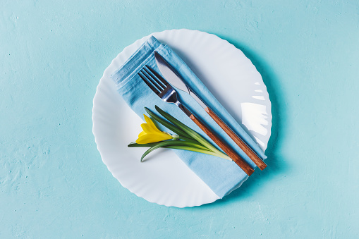 Spring table layout with plate and tableware on pastel blue background. Spring Holidays background. Top view, flat lay, copy space.